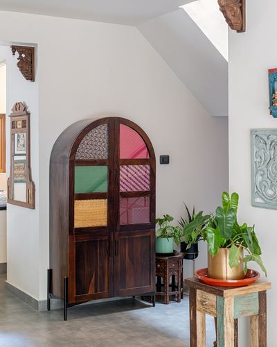 Arched wooden cabinet with patterned fabric panels in a plant-filled foyer with carved wood accents
