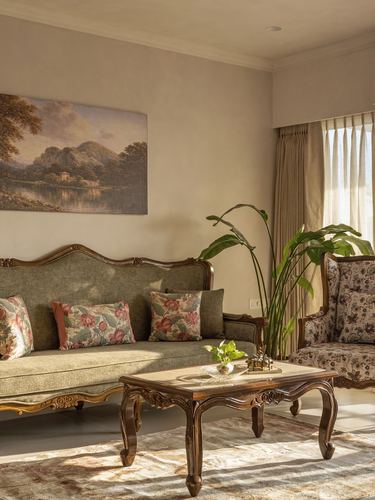 Living room with carved wood sofa, floral armchair, landscape painting, and sunlit curtains