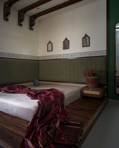 Raised platform bed with green upholstered backrest, carved wood ceiling beams, and maroon silk throw in Chennai home.