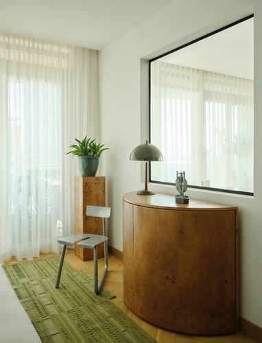 Curved burl wood cabinet under large mirror beside sheer curtained window, striped rug on herringbone floor
