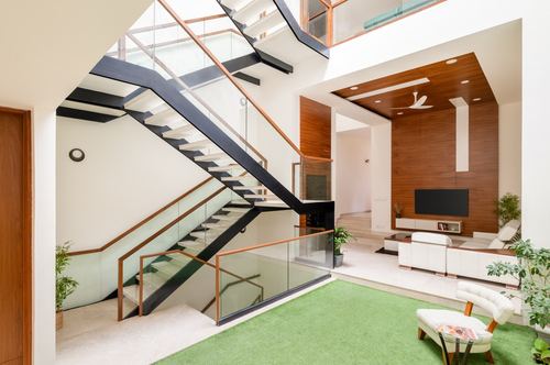 Double-height living room with floating staircase, glass railing, and wood-paneled TV wall beyond a green rug