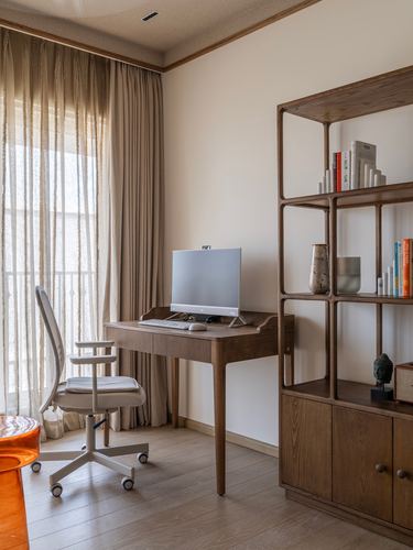 A wooden study desk with monitor beside sheer curtained window and an open wooden shelving unit with books and vases