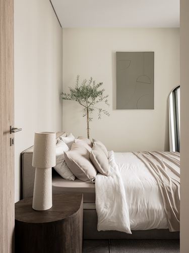 Minimalist neutral bedroom with upholstered bed, textured lamp, round wood table, and abstract print
