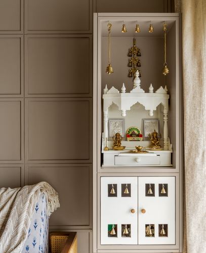 White marble temple-shaped pooja unit with hanging brass bells, set into a taupe paneled wardrobe wall