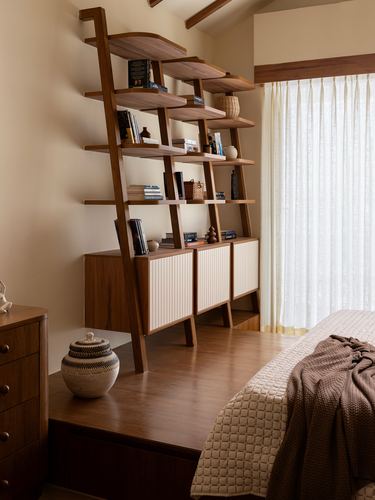 Wooden ladder shelf with fluted cabinets beside a bed and sheer curtained window in a warm-toned bedroom.
