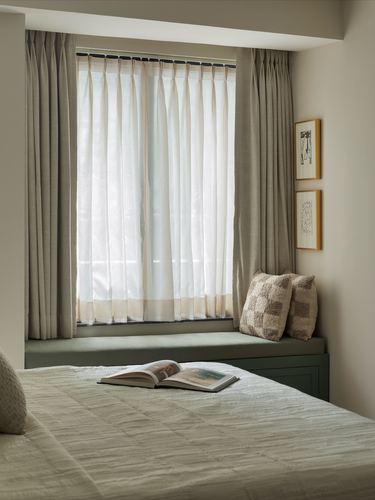 Bedroom with sheer-curtained window seat in sage green, checkered pillows, and bed with open book in foreground