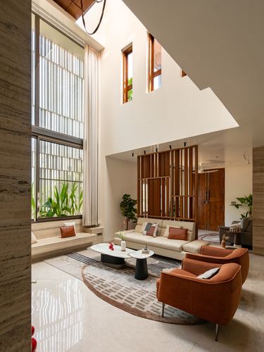 Double-height living room with louvered windows, stone floor, wooden screen and terracotta armchairs