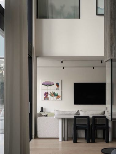 Double-height living room wall with framed art panels, tv, dining table and black chairs seen through a doorway
