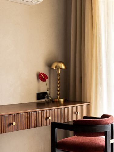Fluted wood console with brass lamp and red flower beside a velvet chair near sheer curtains