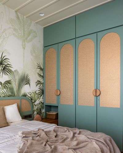 Sage green wardrobe with arched cane door panels beside a bed and palm-leaf wallpaper wall
