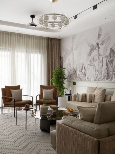 Living room with tan leather armchairs, beige sofas, and a botanical mural wall under a round chandelier