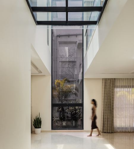 Woman walks through sunlit corridor beside tall glass atrium with plants and carved screen