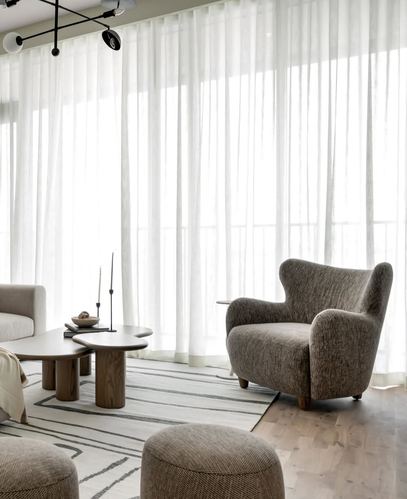 Living room with sheer curtains, grey wingback armchair, and wooden nesting coffee tables on a striped rug