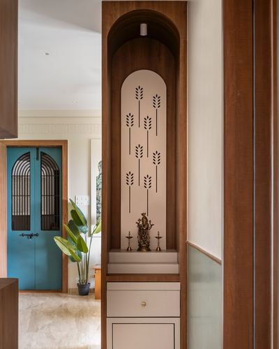 Arched teak pooja niche with carved panel, brass idol, and diyas beside a teal jaali door