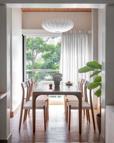 Wooden dining table with woven chairs beneath a paper pendant light facing a green balcony view