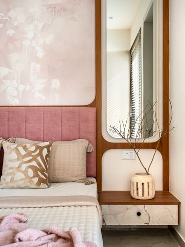 Pink upholstered headboard beside an arched wood-framed mirror and marble bedside ledge with vase.