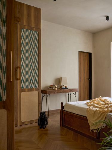 Wooden bedroom with ikat-patterned wardrobe, console table, herringbone floor, and a black cat sitting on the floor