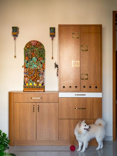 Wooden pooja cabinet with carved deity niche, brass lotus inlays, and a white dog nearby