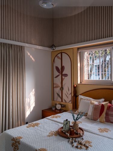 Sunlit bedroom corner with fluted ceiling, painted arched cupboard, and floral bedspread near a window