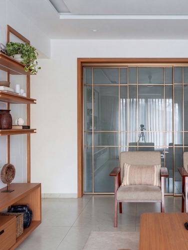 Living room with open wood shelving, cane armchair, and brass-framed glass partition to study beyond