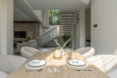 Dining table set for four with staircase and living room visible beyond in sunlit contemporary home