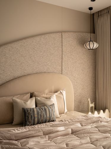 Beige bedroom with curved boucle headboard panel, layered cushions, and a black pendant lamp beside the bed