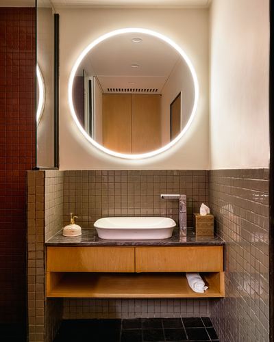 Bathroom vanity with round backlit mirror, olive tile, stone-top wood cabinet, and vessel sink