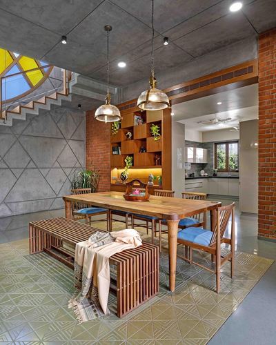 Wooden dining table with blue cushioned chairs and slatted bench under brass pendant lights, brick wall shelving behind