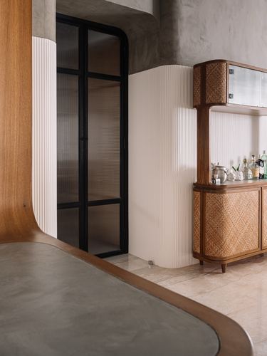 Curved fluted white wall beside black-framed reeded glass door and rattan bar cabinet