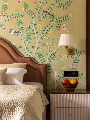Bedroom corner with hand-painted bird and floral mural wallpaper, terracotta headboard, and fluted nightstand with fruit bowl