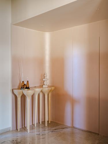 Sculptural wood-legged console table in a warm pink corner niche with diffuser bottles and stone sculpture, marble floor.
