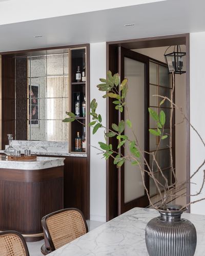 Marble dining table with vase beside a fluted wood bar and mirrored shelving, glass door beyond