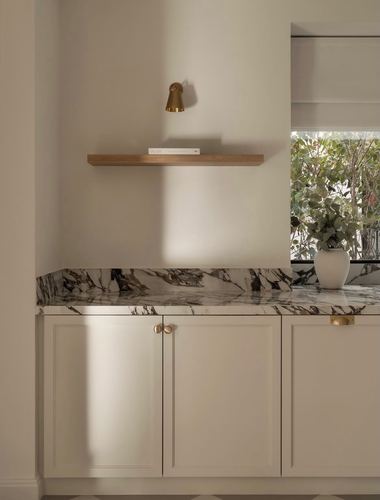 Cream shaker cabinets under a veined marble backsplash with a brass sconce and oak shelf