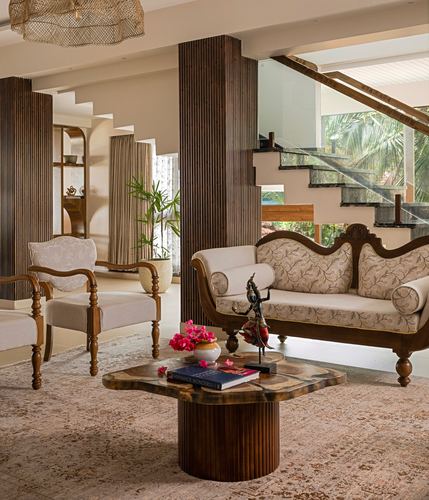 Living room with carved wooden settee, fluted columns, and staircase framed by greenery beyond