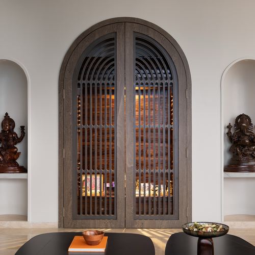 Arched fluted wooden door opens to a glowing pooja niche, flanked by carved deity statues in wall alcoves