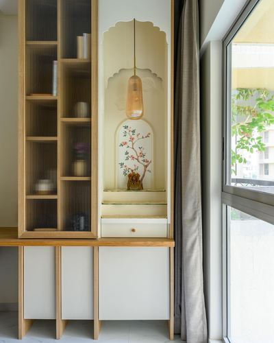 Arched pooja niche with floral mural and pendant light built into wooden cabinetry beside window