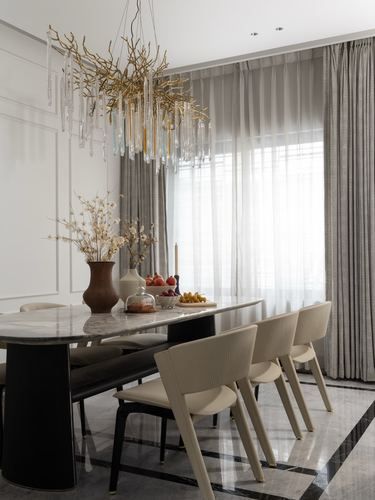 Dining room with marble table, cream chairs, and a branch-form crystal chandelier lit by sheer curtained window