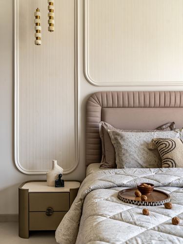Taupe channel-tufted headboard beside curved fluted wall panel and brass drawer nightstand with pendant lights