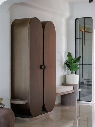 Rounded bronze-toned wardrobe beside bench and plant in a marble-floored foyer