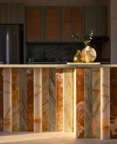 Kitchen island clad in vertical onyx and stone strips, backlit by afternoon sun, arched cane cabinets behind