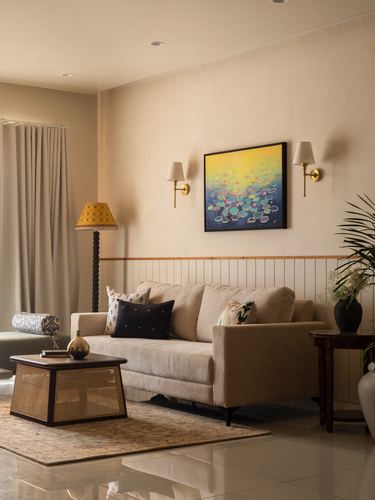Beige living room with cane coffee table, sofa, brass sconces and colorful framed artwork above wainscoting