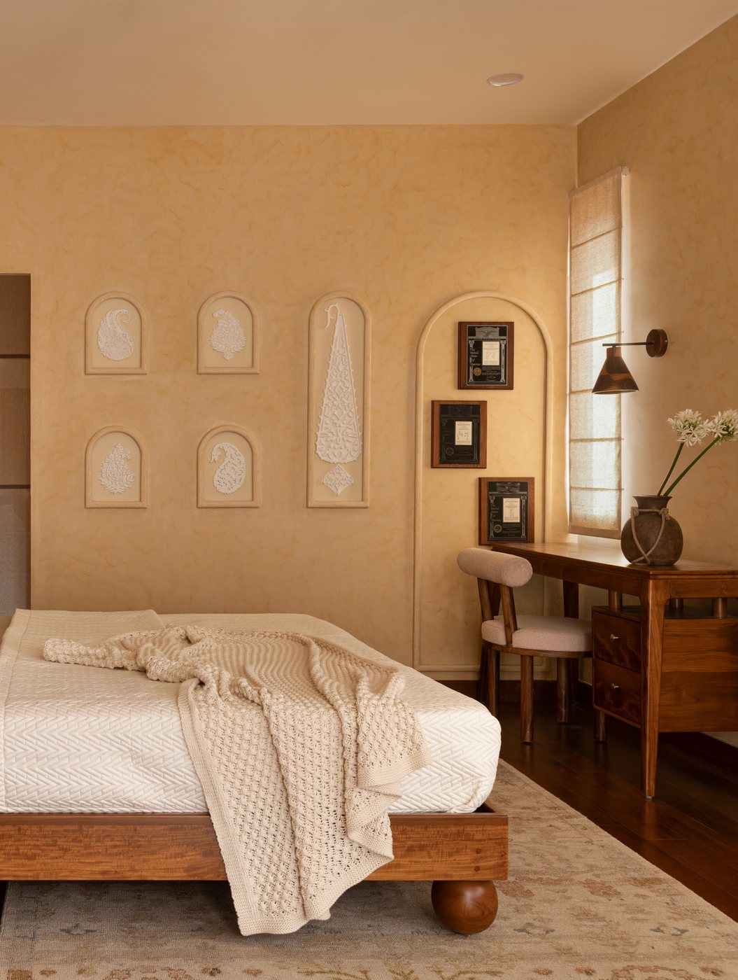 Bedroom with limewashed walls, arched carved niches, wooden bed and desk lit by soft daylight