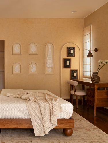 Bedroom with limewashed walls, arched carved niches, wooden bed and desk lit by soft daylight