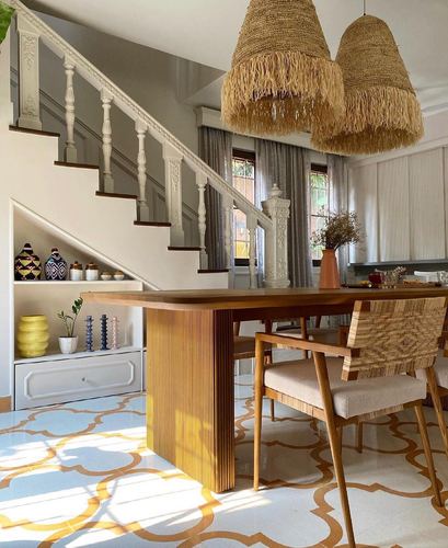 Dining table with cane chairs under fringed raffia pendants beside a carved staircase on patterned tile floor