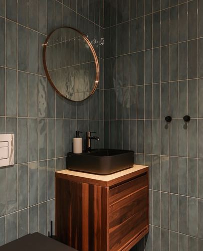 Corner bathroom vanity with round brass mirror against teal vertical tile and walnut cabinet