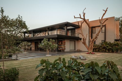 Two-storey earthy-toned villa with dark cantilevered overhang, glazing, and landscaped garden with sculptural tree