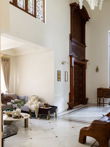 Double-height foyer with ornately carved wooden door, marble inlay floor, and leather stool in foreground