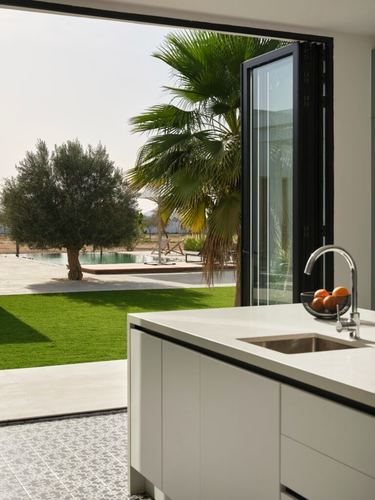 White kitchen island beside open bifold glass doors overlooking lawn, pool, and palm trees