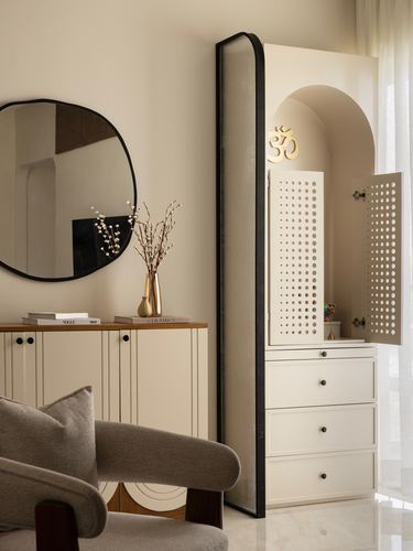 Arched pooja unit with perforated jali doors beside a curved mirror and cane cabinet in a neutral room