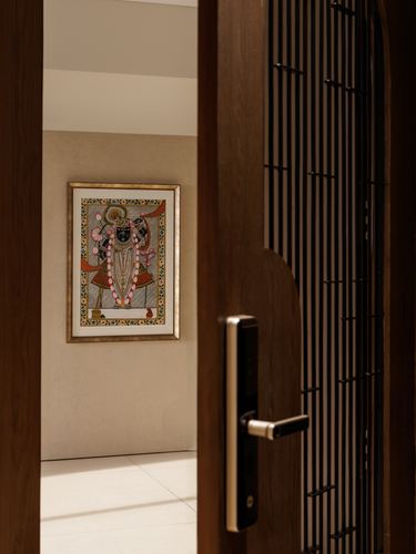 Open wooden door reveals a framed pichwai painting on a beige wall down a hallway
